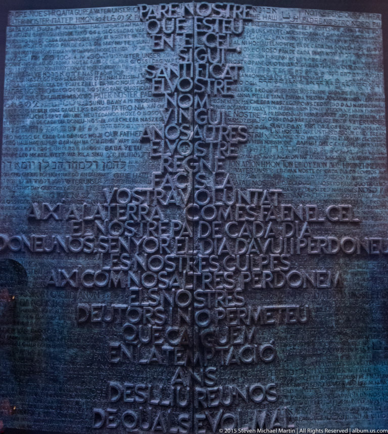 Dark metal Sagrada Família door densely covered with raised multilingual text from the Lord’s Prayer.
