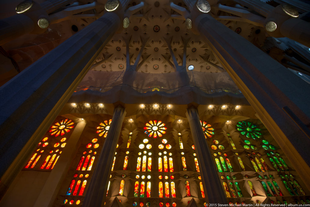 Red, orange, and yellow stained-glass windows glowing below branching columns inside the dark Sagrada Família nave.