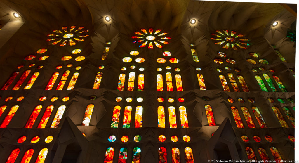Wall of red, orange, yellow, and green Sagrada Família stained glass with rosette forms framed by branching stone supports.