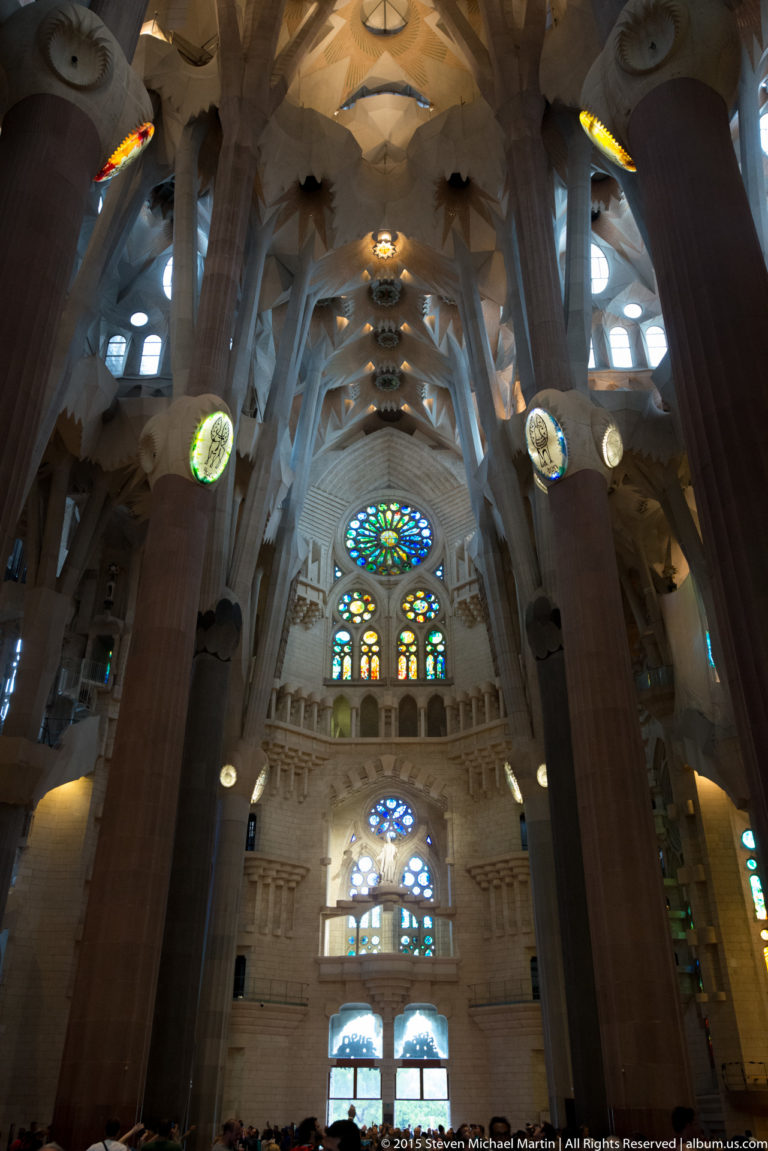Sagrada Família interior with massive columns framing a tall wall of blue stained glass and circular windows.