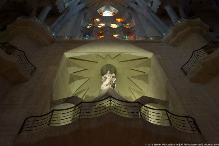 Pale religious statue in a starburst-shaped niche above a curved gallery, with stained-glass openings overhead.