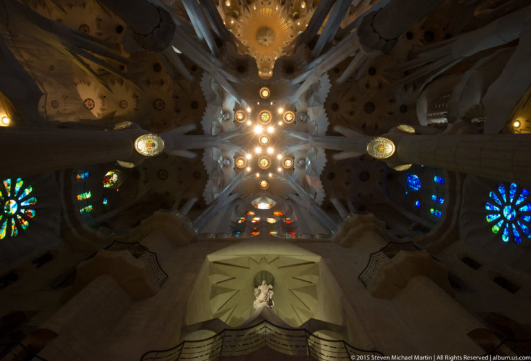 Upward Sagrada Família interior showing branching vault, circular lights, stained glass, and a small statue in a geometric niche.