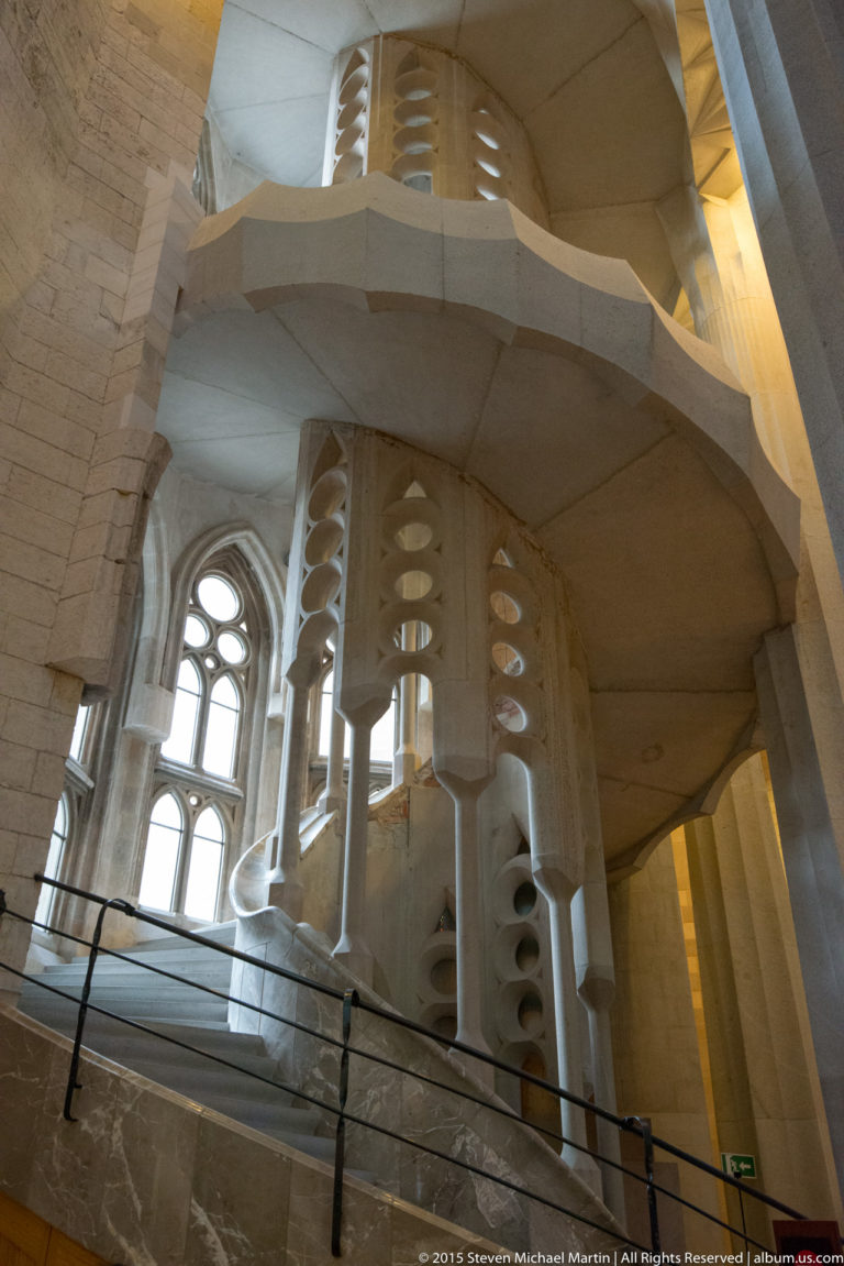 Curving pale stone galleries with vertical supports pierced by repeated oval openings inside the Sagrada Família.