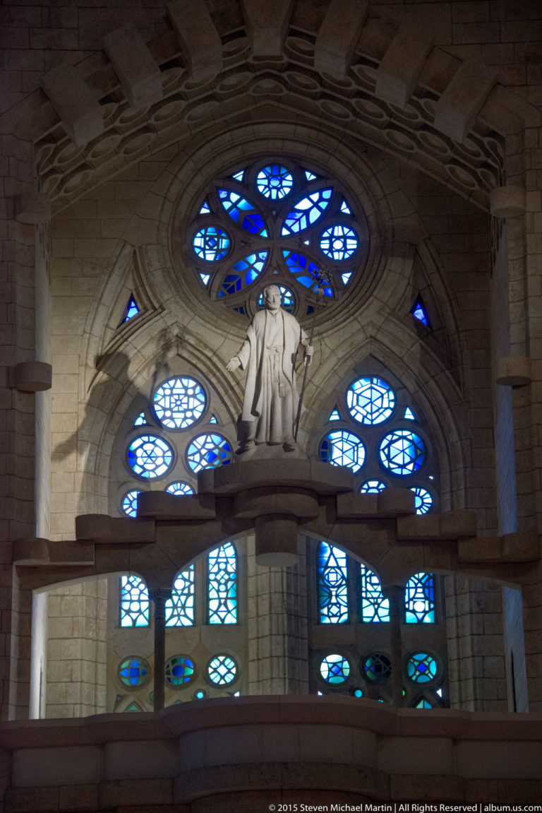 Pale standing statue on an elevated ledge before blue circular and geometric stained-glass windows inside the Sagrada Família.