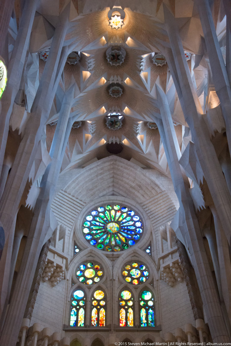 Upward interior view with a large multicolored circular window, smaller stained glass, branching columns, and illuminated vaults.