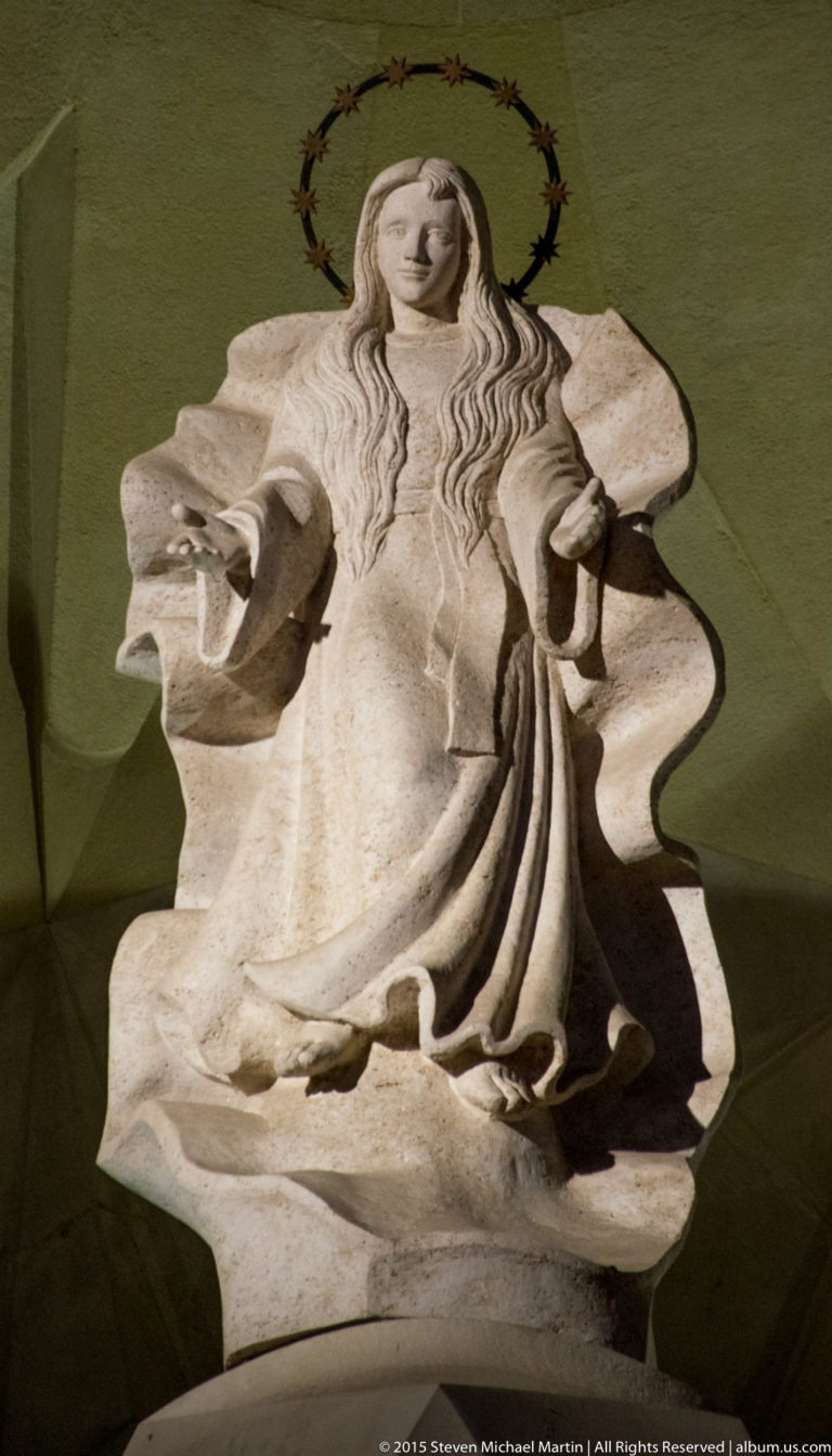 Stone figure of the Virgin Mary in flowing robes with a circular thorn-like halo against a green wall.