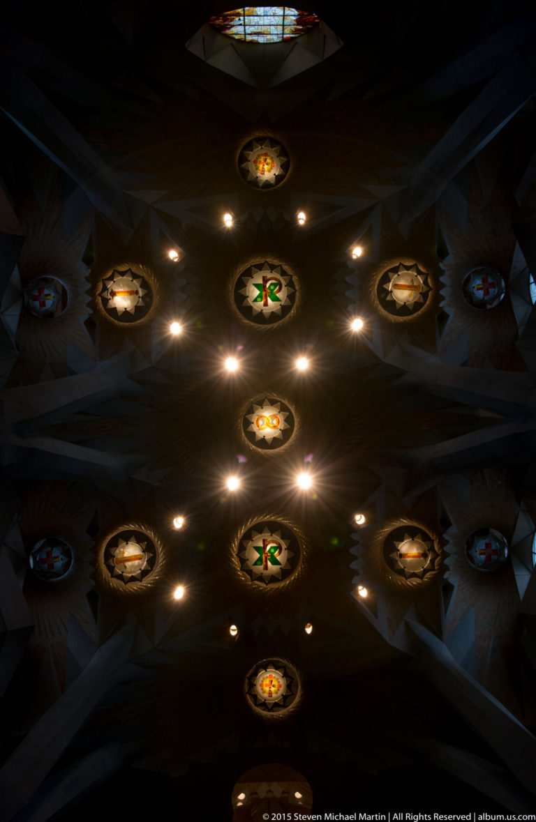 Dark Sagrada Família ceiling with a line of illuminated circular symbolic medallions and small lights between branching supports.