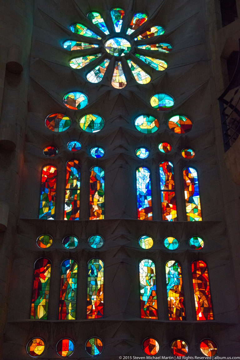 Tall Sagrada Família wall filled with green, blue, red, and orange stained-glass windows and circular rosette forms.