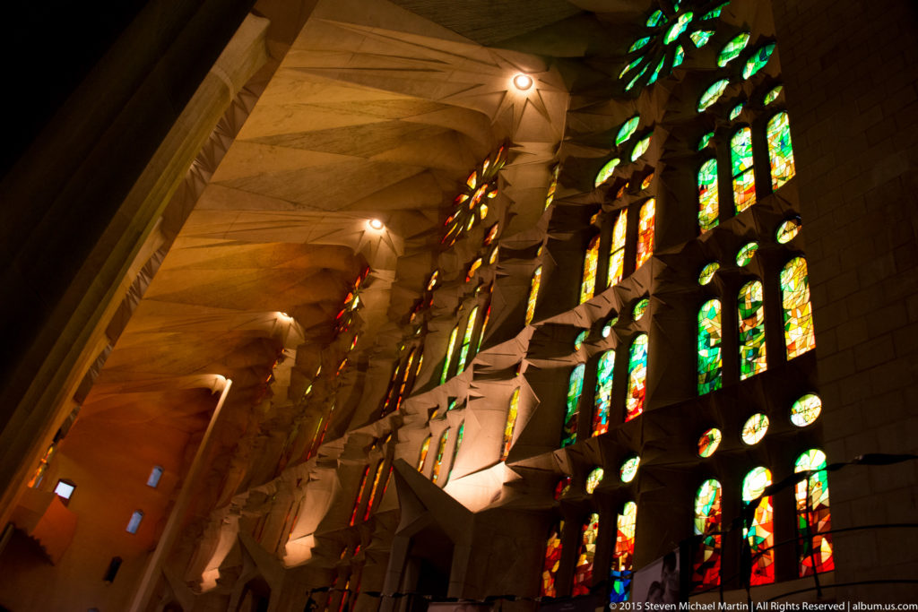 Oblique interior view along multicolored stained-glass windows and angular stone supports in a Sagrada Família aisle.
