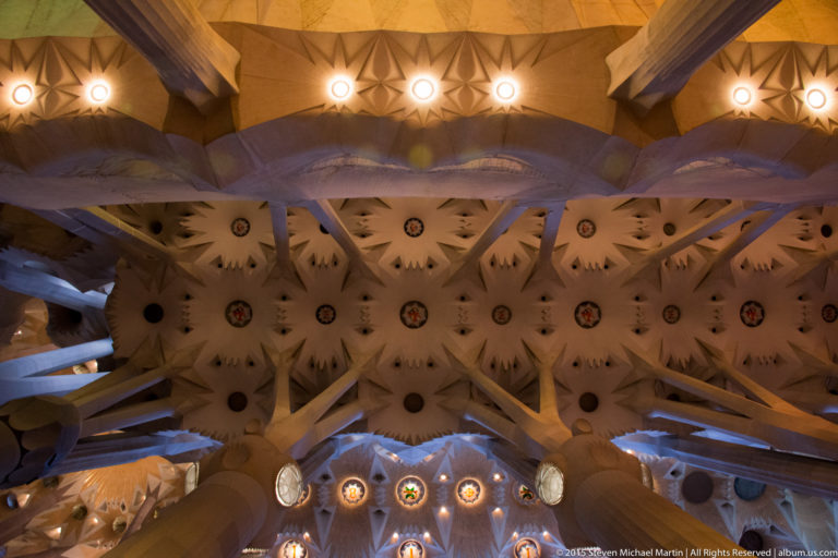 Upward view of star-like geometric vault panels, circular bosses, lights, and branching supports inside the Sagrada Família.