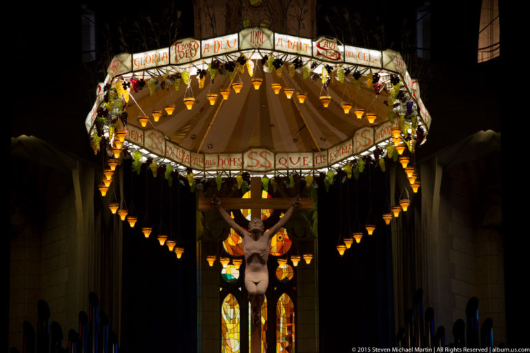 Frontal view of the crucified Christ suspended beneath an illuminated canopy with hanging lamps and stained glass behind.