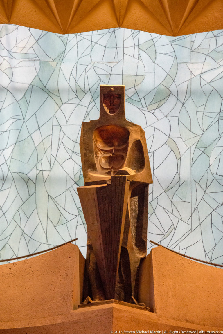 Angular brown liturgical furnishing with stacked geometric forms positioned before pale abstract glazing.