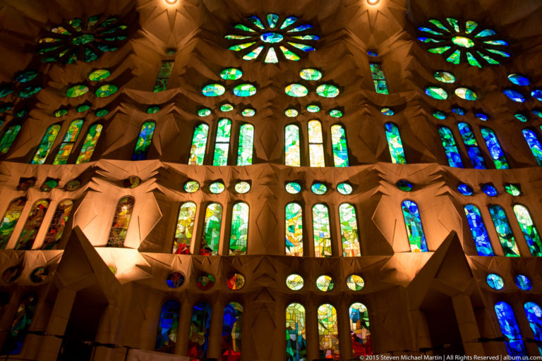 Broad Sagrada Família wall of predominantly green and blue stained glass with circular rosettes and narrow vertical windows.