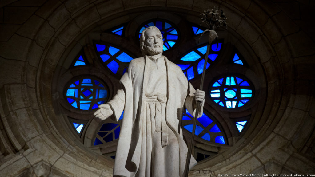 Bearded robed stone figure holding a staff before a bright blue circular stained-glass window.