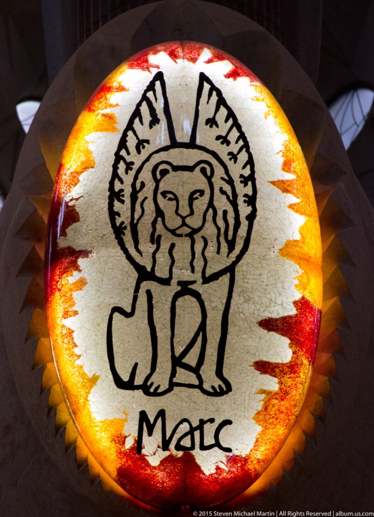 Orange-red circular medallion with a stylized winged lion and the Catalan name Marc.