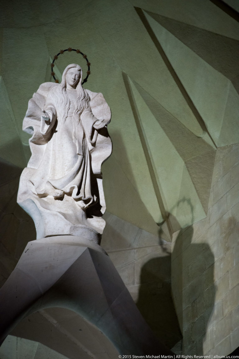 Small pale robed religious statue on a ledge casting a long angular shadow across a greenish wall.
