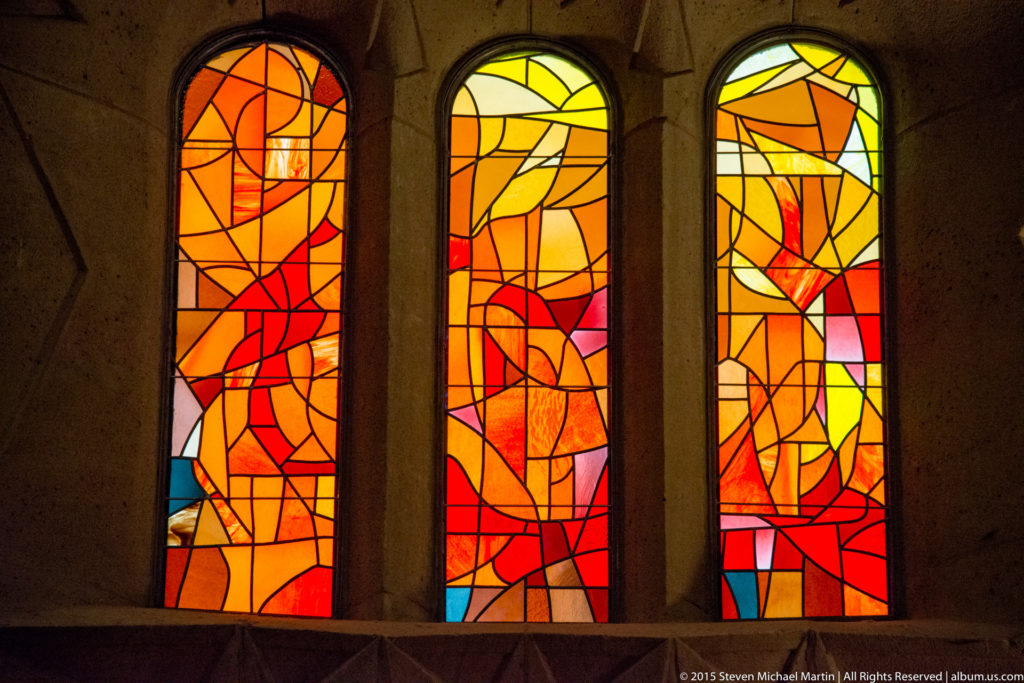 Three tall abstract stained-glass windows in orange, red, yellow, and small blue sections inside the Sagrada Família.