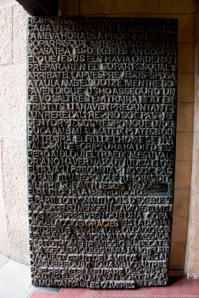 Large dark metal door densely covered with raised words and phrases in varied sizes at the Sagrada Família.