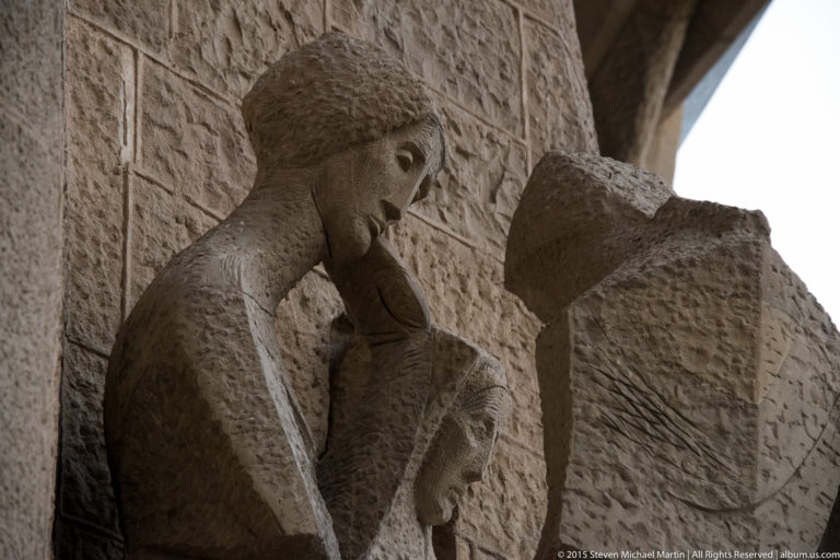 Two angular stone figures leaning toward each other within a recessed sculptural group on the Sagrada Família façade.