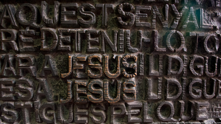 Close view of dark raised capital-letter inscriptions on the Sagrada Família façade, with JESUS repeated across the surface.