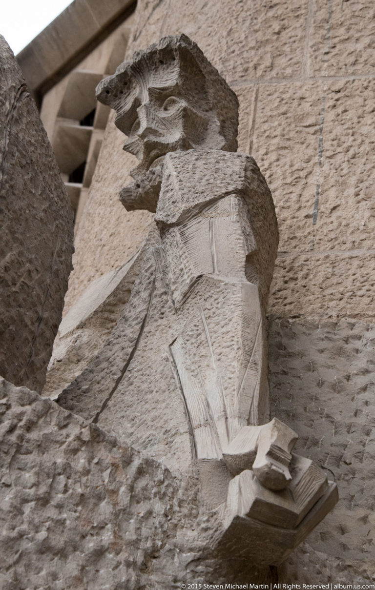 Low-angle view of an angular robed stone figure holding a book on the Sagrada Família Passion façade.
