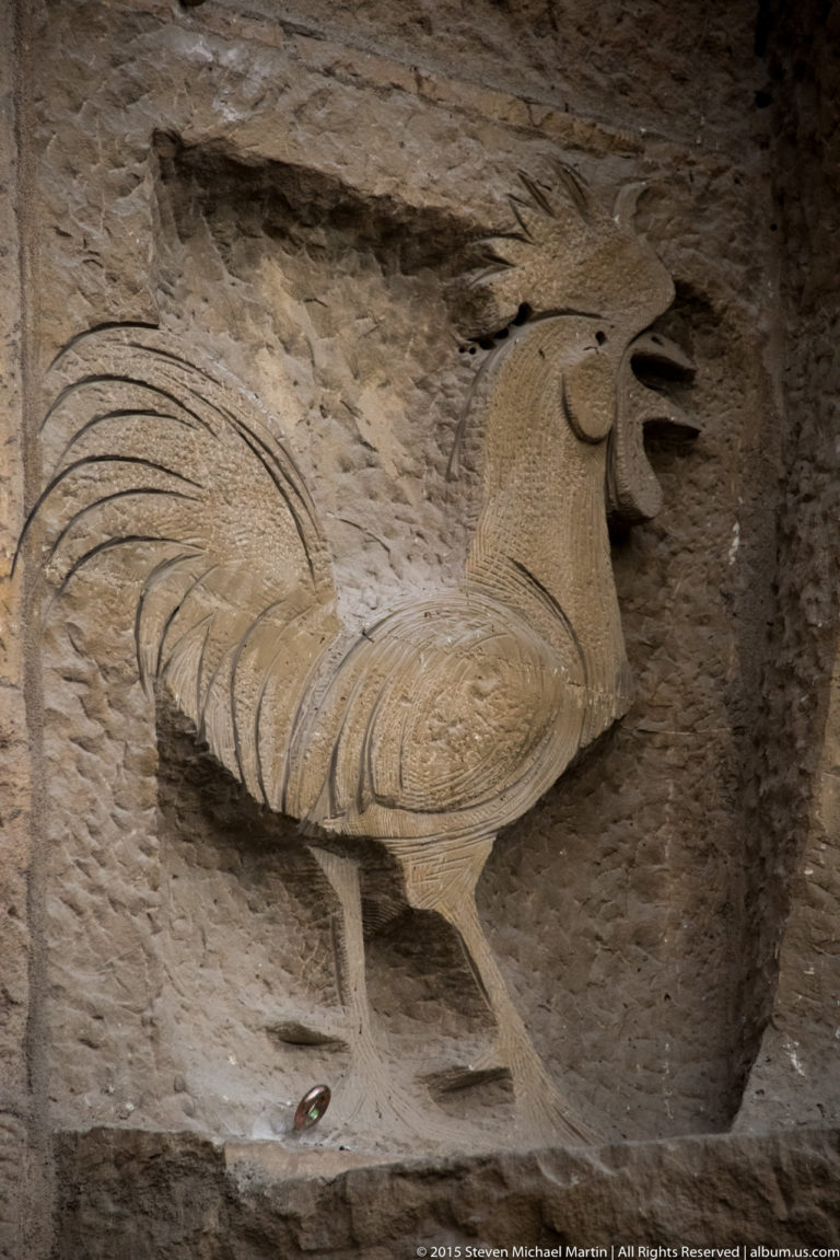 Carved rooster from Peter’s Denial on the Sagrada Família Passion façade, with open beak and long arcing tail.