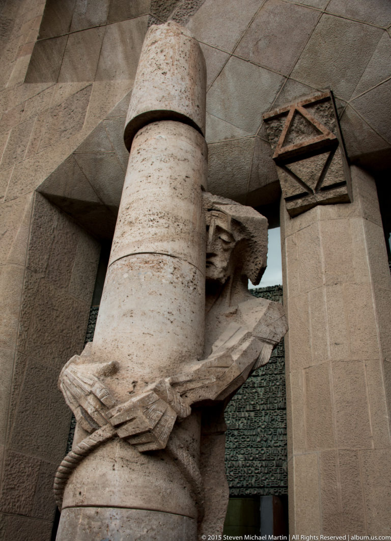 Large cylindrical stone column beside angular carved forms and a dark geometric symbol on the Sagrada Família Passion façade.