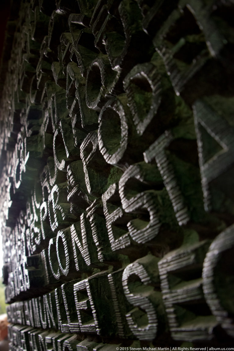 Oblique close view of a dark metal Sagrada Família door densely covered with raised words and oversized letter fragments.
