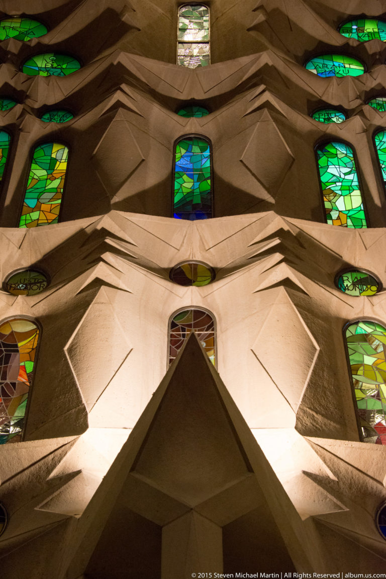 Symmetrical pale geometric wall with projecting angular forms and small green and blue stained-glass windows.