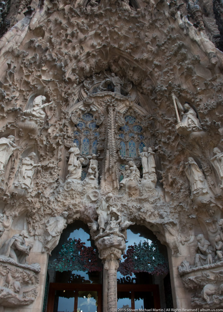 Nativity façade portal densely covered with figures, foliage, textured stone carving, and blue-green glazed openings.