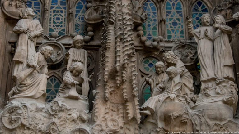 Groups of kneeling, seated, and standing stone figures around a textured column and blue-green windows on the Nativity façade.