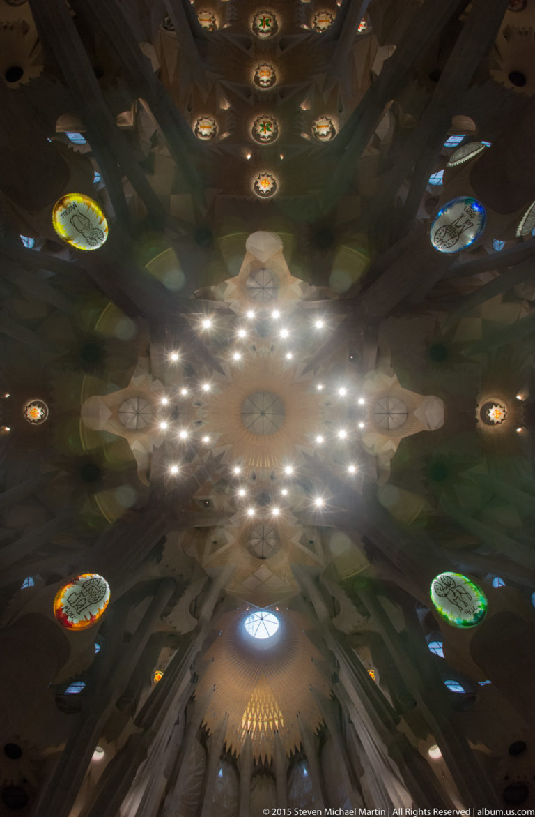 Near-vertical view of the Sagrada Família ceiling with a bright star-like cluster of lights, branching structure, and colored round windows.