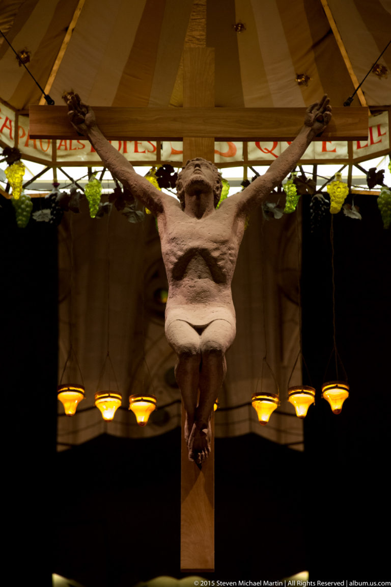 Frontal close view of Christ on the suspended crucifix beneath the Sagrada Família canopy and hanging lamps.