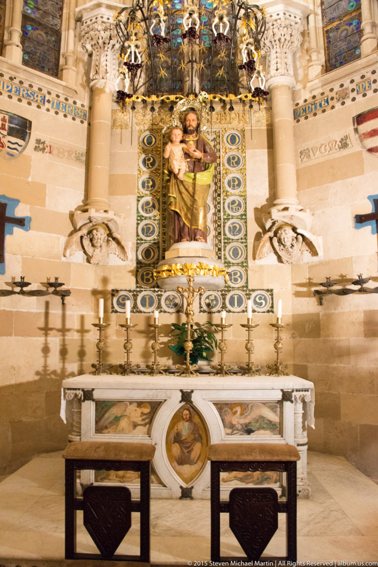 Ornate side altar with a robed male saint holding a child above candles and a decorated altar table.