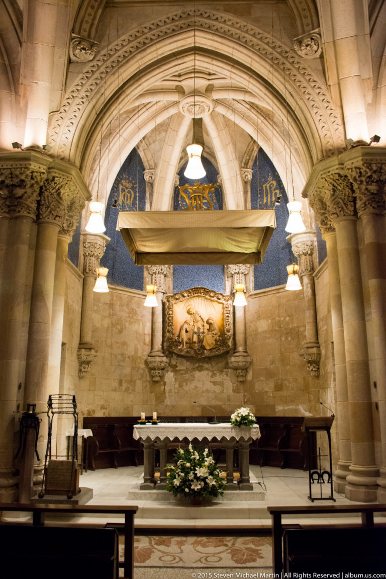 Stone altar beneath an arch with columns, hanging lamps, flowers, blue decoration, and a relief on the rear wall.