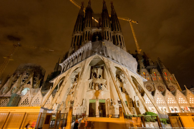 Night view of the illuminated Sagrada Família Passion façade, four bell towers, inclined columns, and construction cranes.