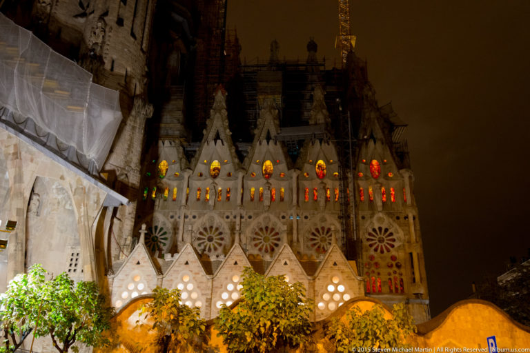 Night exterior of the Sagrada Família with glowing multicolored windows, triangular gables, scaffolding, and a crane.