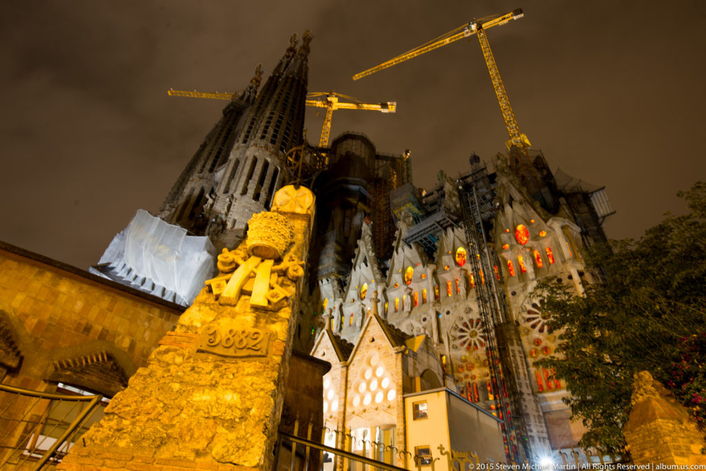 Illuminated foreground sculpture below the Sagrada Família’s towers, glowing windows, and construction cranes at night.