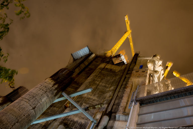 Low-angle nighttime view of unfinished Sagrada Família masonry, temporary supports, platforms, and yellow construction cranes.