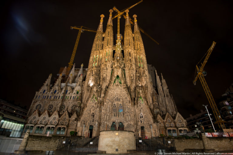 Frontal night view of the illuminated Sagrada Família Nativity façade and four towers, with cranes on both sides.