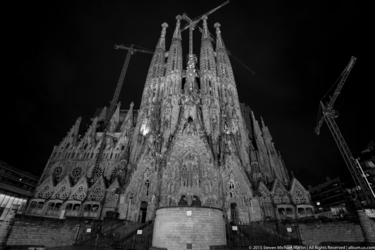 Black-and-white nighttime view of the Sagrada Família Nativity façade, four illuminated towers, and surrounding cranes.