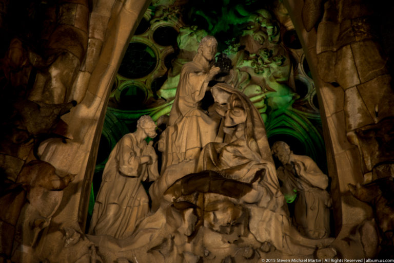 Night-lit Coronation of Mary on the Nativity façade, with Christ crowning the Virgin as Saint Joseph and a shepherd look on.