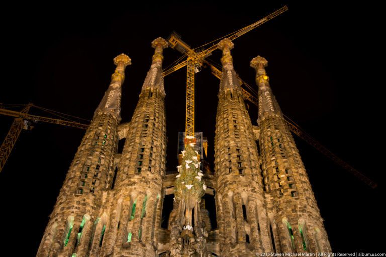 Four illuminated Sagrada Família bell towers rising against a black night sky with construction cranes behind them.