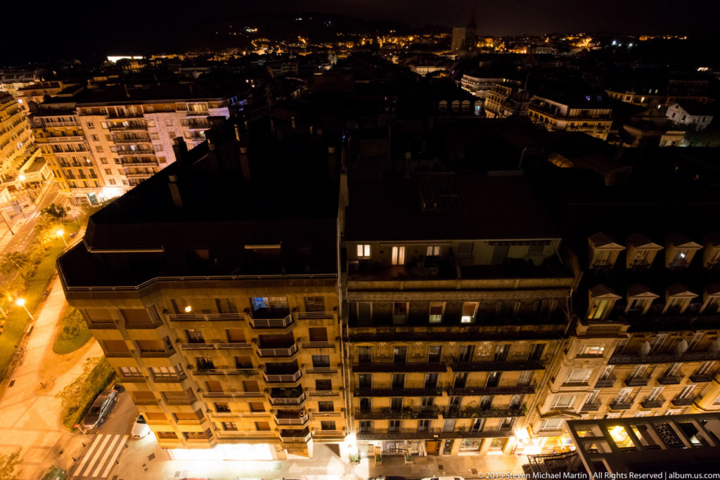 Elevated nighttime view across apartment rooftops and illuminated streets in central San Sebastián, with dark hills beyond.