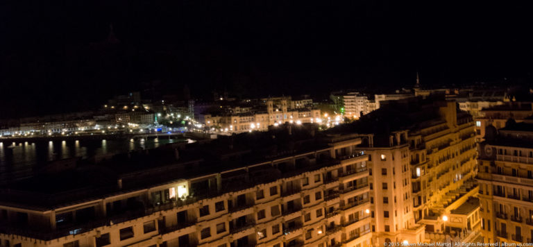 Night view over foreground rooftops toward San Sebastián’s brightly illuminated waterfront and dark bay.