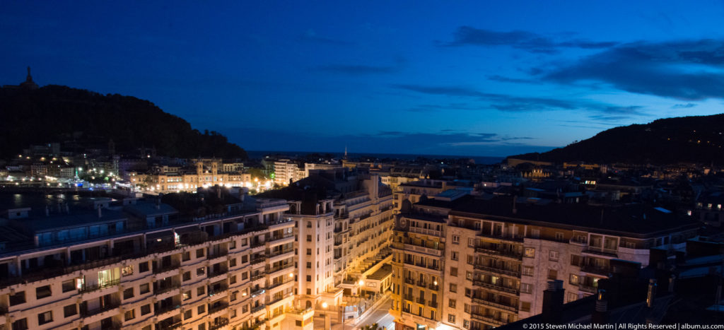 Blue-hour view across illuminated San Sebastián apartment blocks toward the sea between dark hills.
