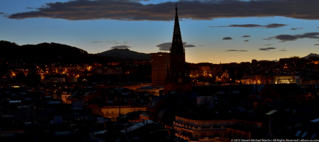 Silhouetted pointed church spire centered above dark San Sebastián rooftops at twilight.