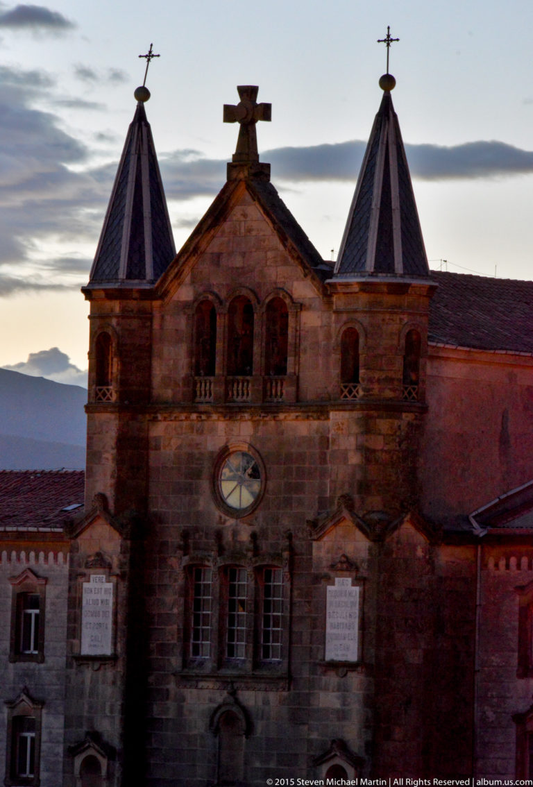 Historic masonry façade with twin pointed-roof towers, crosses, arched openings, and a central clock at dusk.