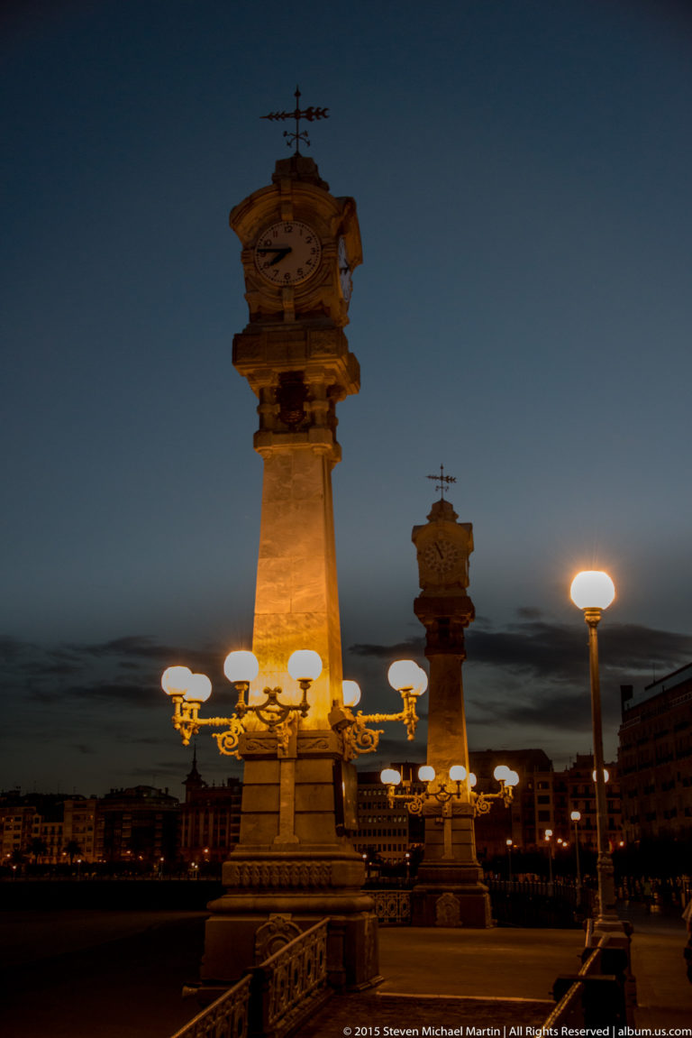 Illuminated ornamental clock column and globe lamps on San Sebastián’s seafront promenade at dusk.