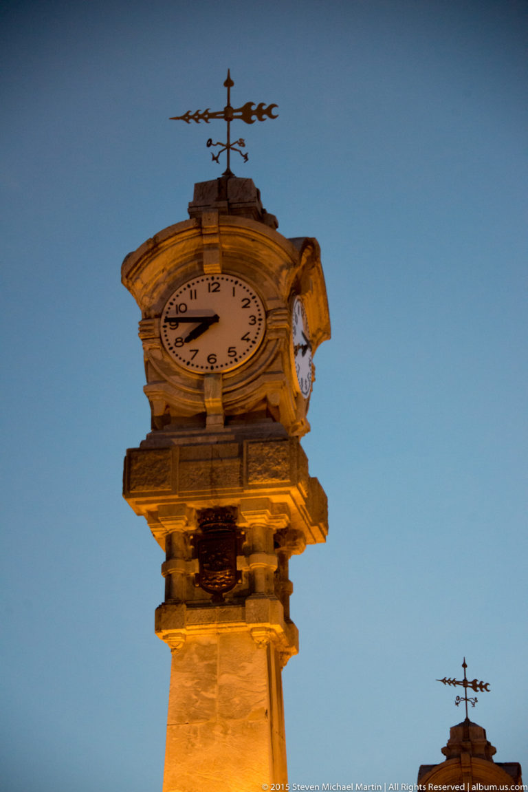 Close-up of an illuminated four-faced stone clock with weather vane against a blue evening sky.